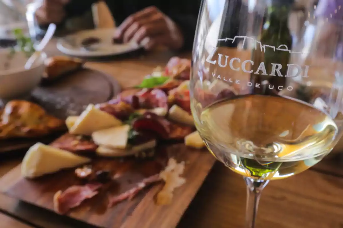 Copa de vino blanco con la marca Zuccardi en primer plano y una tabla de quesos y fiambres desenfocada al fondo sobre una mesa de madera Copa de vino blanco con la marca Zuccardi en primer plano y una tabla de quesos y fiambres desenfocada al fondo sobre una mesa de madera