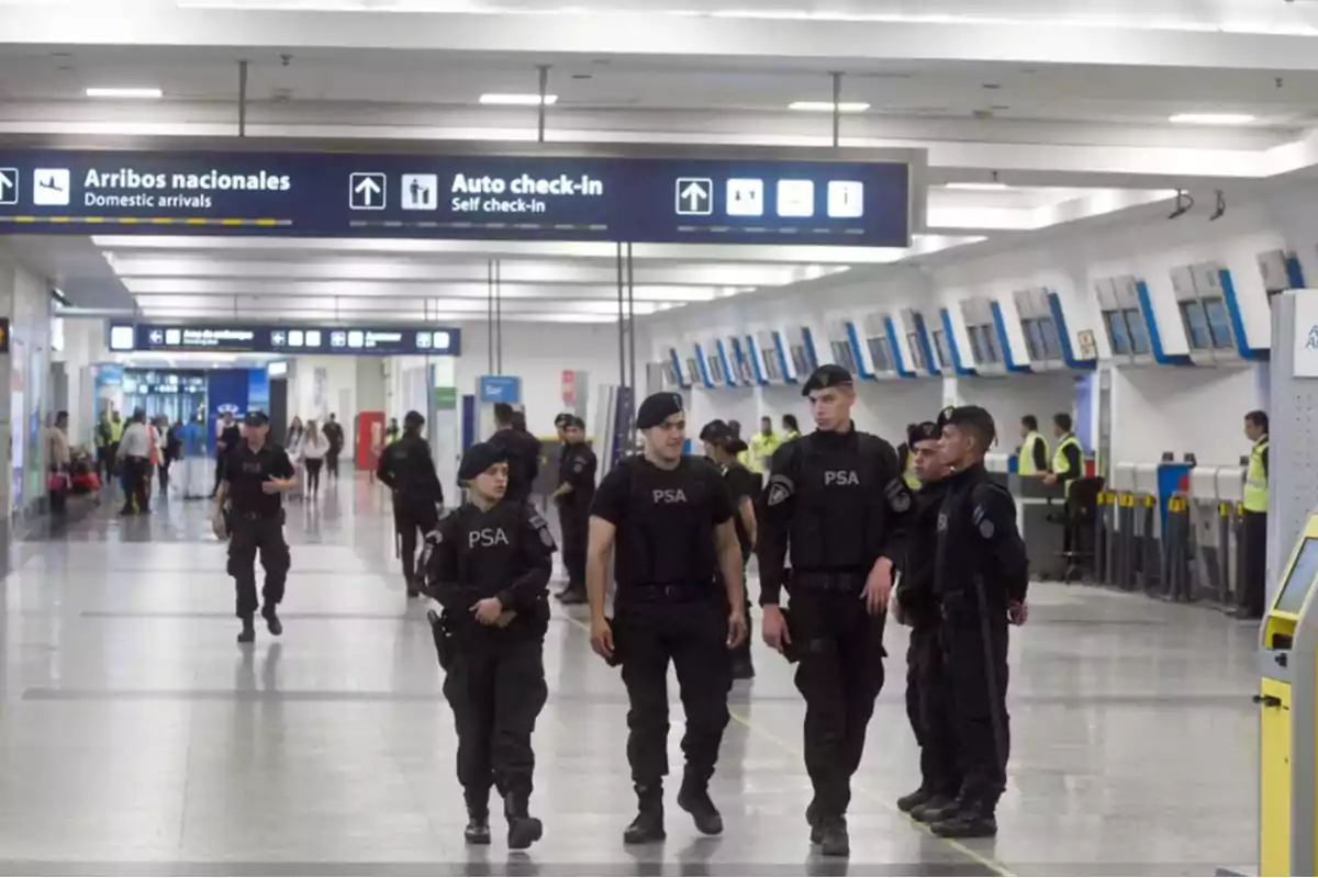 Varios agentes de seguridad vestidos de negro patrullan el área de check-in en un aeropuerto mientras algunas personas caminan al fondo Varios agentes de seguridad vestidos de negro patrullan el área de check-in en un aeropuerto mientras algunas personas caminan al fondo