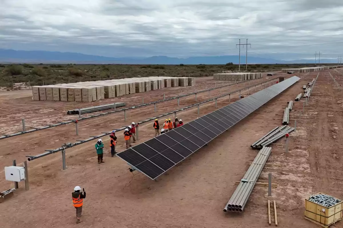 People wearing vests and helmets watch the installation of solar panels on a wide, desert-like area with construction materials stacked around. People wearing vests and helmets watch the installation of solar panels on a wide, desert-like area with construction materials stacked around.