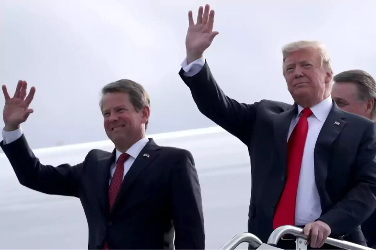 Two men in dark suits and red ties wave with raised hands while standing next to an airplane staircase.