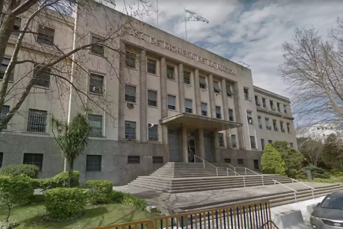 Facade of an institutional building with an access staircase, columns, and a flag waving on the roof, surrounded by trees and gardens.