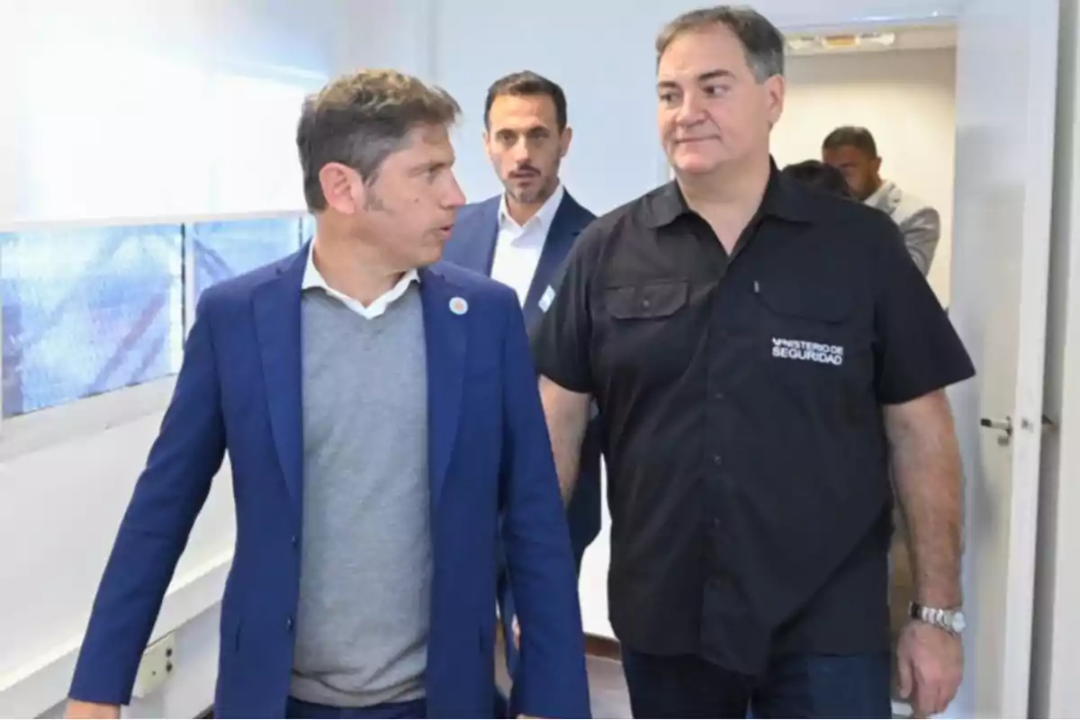 Three men walk down a hallway talking; one of them is wearing a blue jacket and another is wearing a black jersey with the words Ministry of Security.