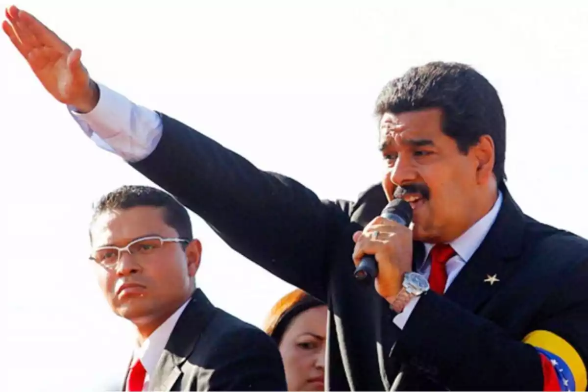 Man in a dark suit with a microphone raising his right arm while another person watches him Man in a dark suit with a microphone raising his right arm while another person watches him
