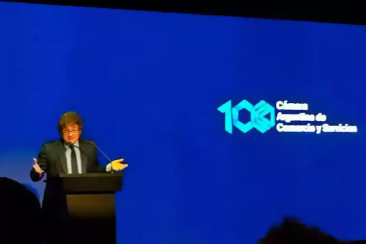 Man in a suit speaking at a podium in front of a blue screen with the logo of the Argentine Chamber of Commerce and Services celebrating 100 years. Man in a suit speaking at a podium in front of a blue screen with the logo of the Argentine Chamber of Commerce and Services celebrating 100 years.