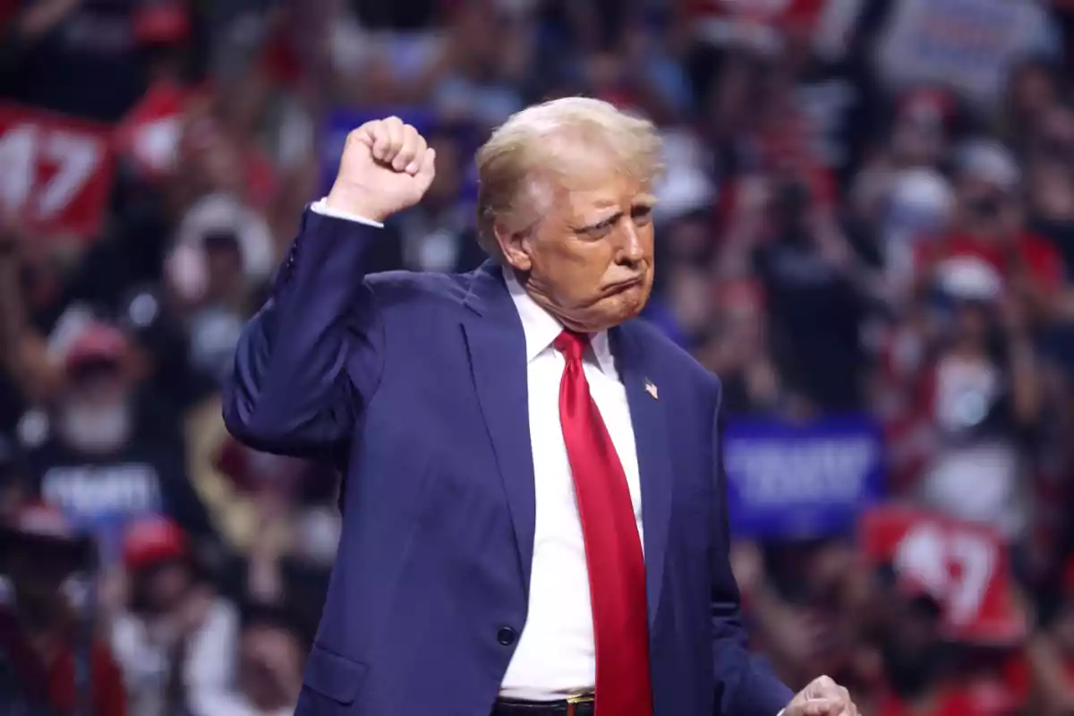 Blond-haired man in a blue suit and red tie raising his fist at a crowded event