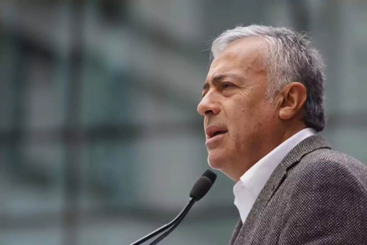 Gray-haired man speaking in front of a microphone with a blurred building in the background
