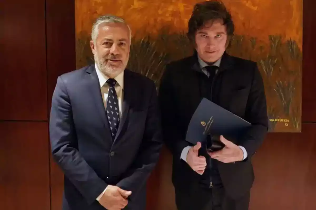 Two men in suits pose together in front of a painting with an orange and brown background, one of them holding a blue folder. Two men in suits pose together in front of a painting with an orange and brown background, one of them holding a blue folder.