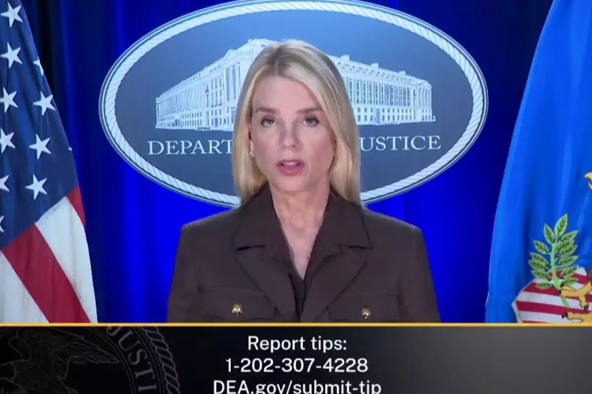 Mujer rubia de cabello lacio hablando frente a un podio con el logo del Departamento de Justicia de fondo y banderas a los lados