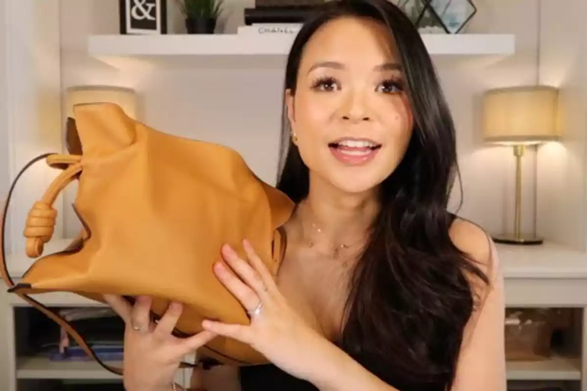 Mujer sonriente mostrando un bolso color camel en una habitación con estantería y lámparas de fondo