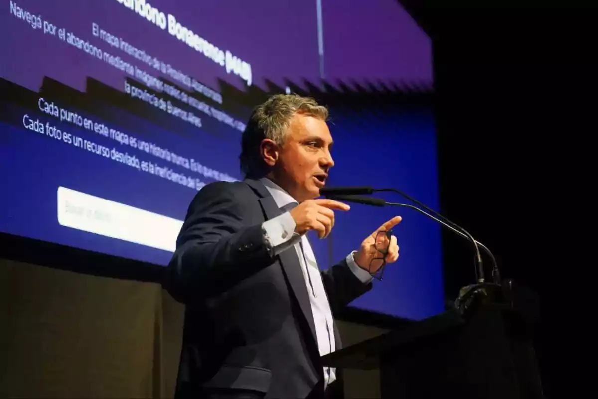 Hombre de cabello canoso hablando en un podio con micrófonos frente a una pantalla grande con texto en un evento o conferencia Hombre de cabello canoso hablando en un podio con micrófonos frente a una pantalla grande con texto en un evento o conferencia