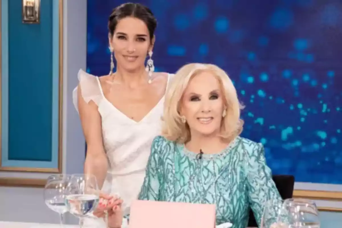 Two elegantly dressed women pose on a television set with glasses of water on the table.
