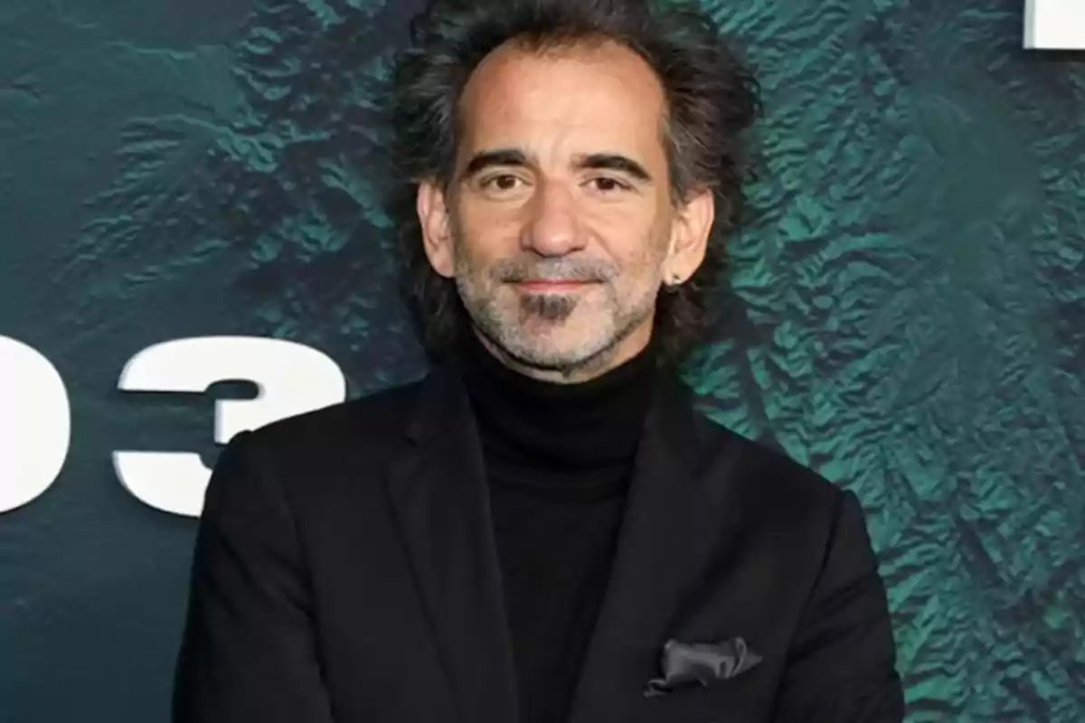 Hombre de cabello oscuro y canoso con barba y bigote, vestido con un saco negro y suéter de cuello alto, posando frente a un fondo verde con letras blancas grandes Hombre de cabello oscuro y canoso con barba y bigote, vestido con un saco negro y suéter de cuello alto, posando frente a un fondo verde con letras blancas grandes