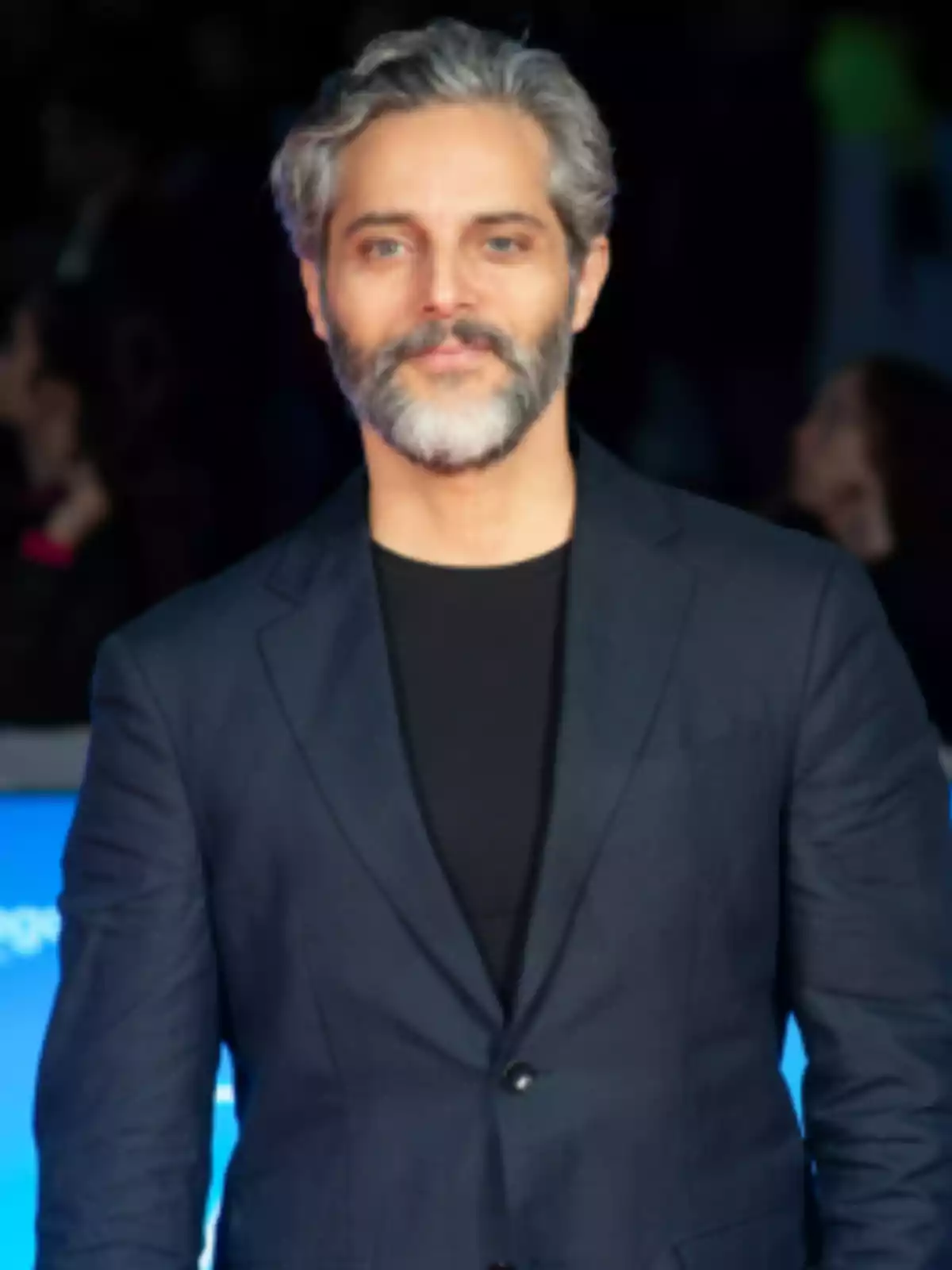 Gray-haired, bearded man wearing a dark blue blazer and a black t-jersey at a formal event Gray-haired, bearded man wearing a dark blue blazer and a black t-jersey at a formal event