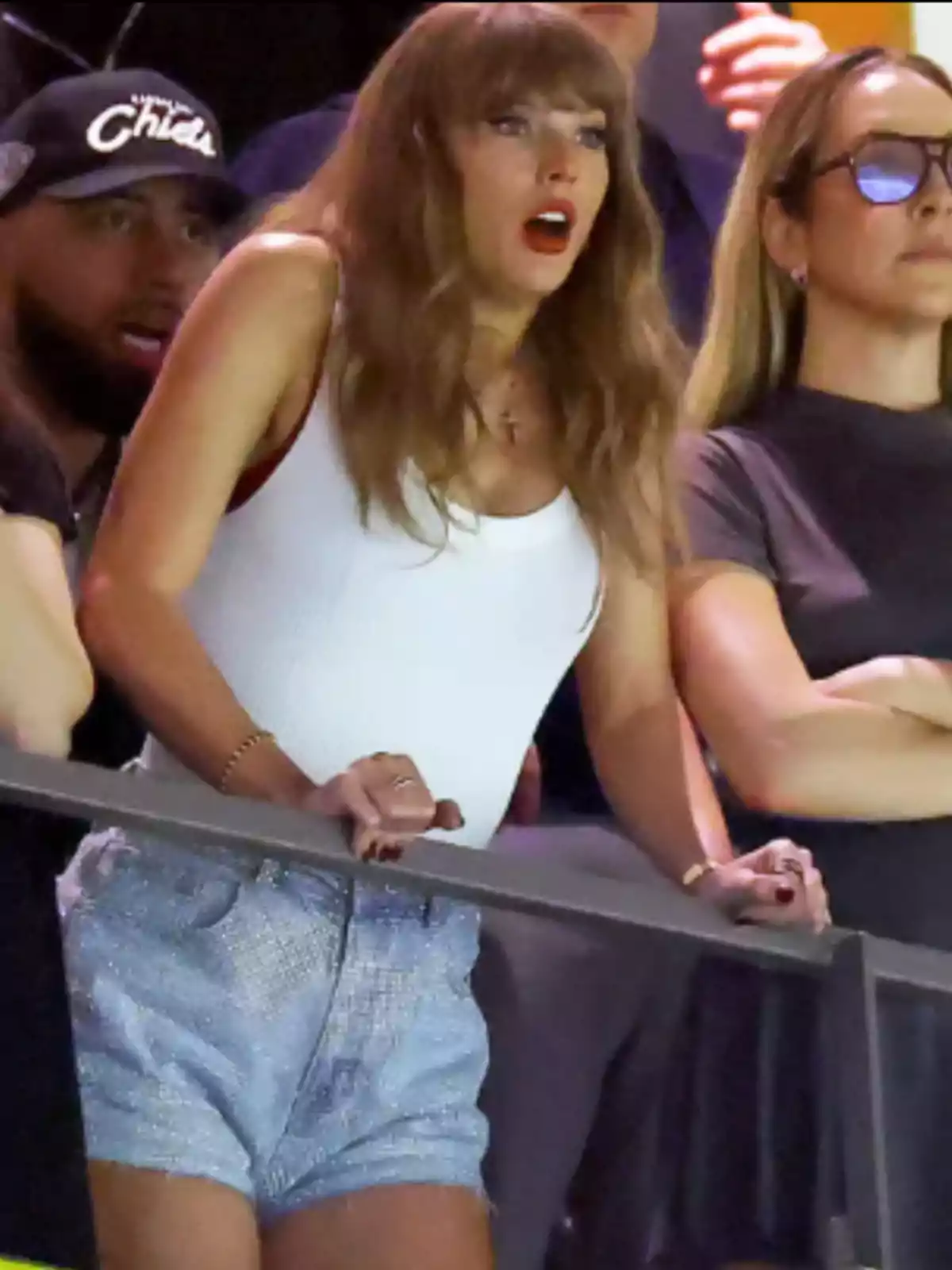 Woman with a surprised expression wearing a white t-jersey and denim shorts at a sporting event surrounded by other people Woman with a surprised expression wearing a white t-jersey and denim shorts at a sporting event surrounded by other people