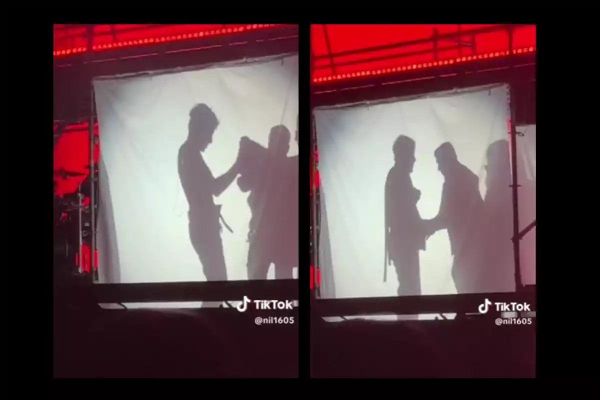 Two people cast their shadows on a white cloth lit with red light while one of them appears to be helping the other put on a garment. Two people cast their shadows on a white cloth lit with red light while one of them appears to be helping the other put on a garment.
