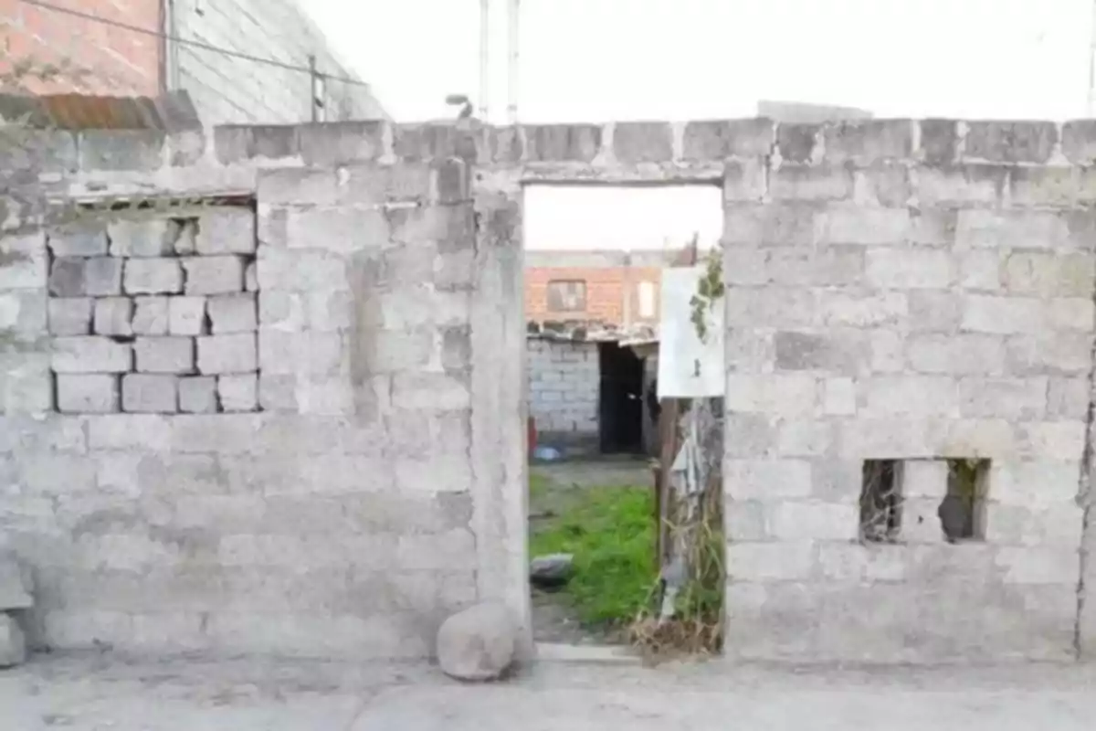 Cement block wall with a frameless door and improvised windows, one of them covered with more blocks and another with branches; in the background, there is a yard with grass and another similar structure. Cement block wall with a frameless door and improvised windows, one of them covered with more blocks and another with branches; in the background, there is a yard with grass and another similar structure.