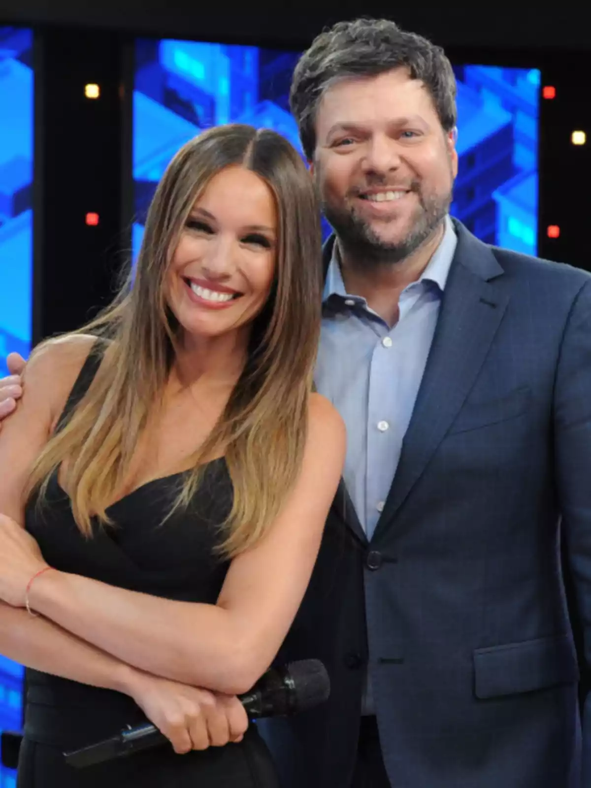 Dos personas sonríen frente a la cámara en un set de televisión, una de ellas sostiene un micrófono y ambos visten ropa elegante Dos personas sonríen frente a la cámara en un set de televisión, una de ellas sostiene un micrófono y ambos visten ropa elegante