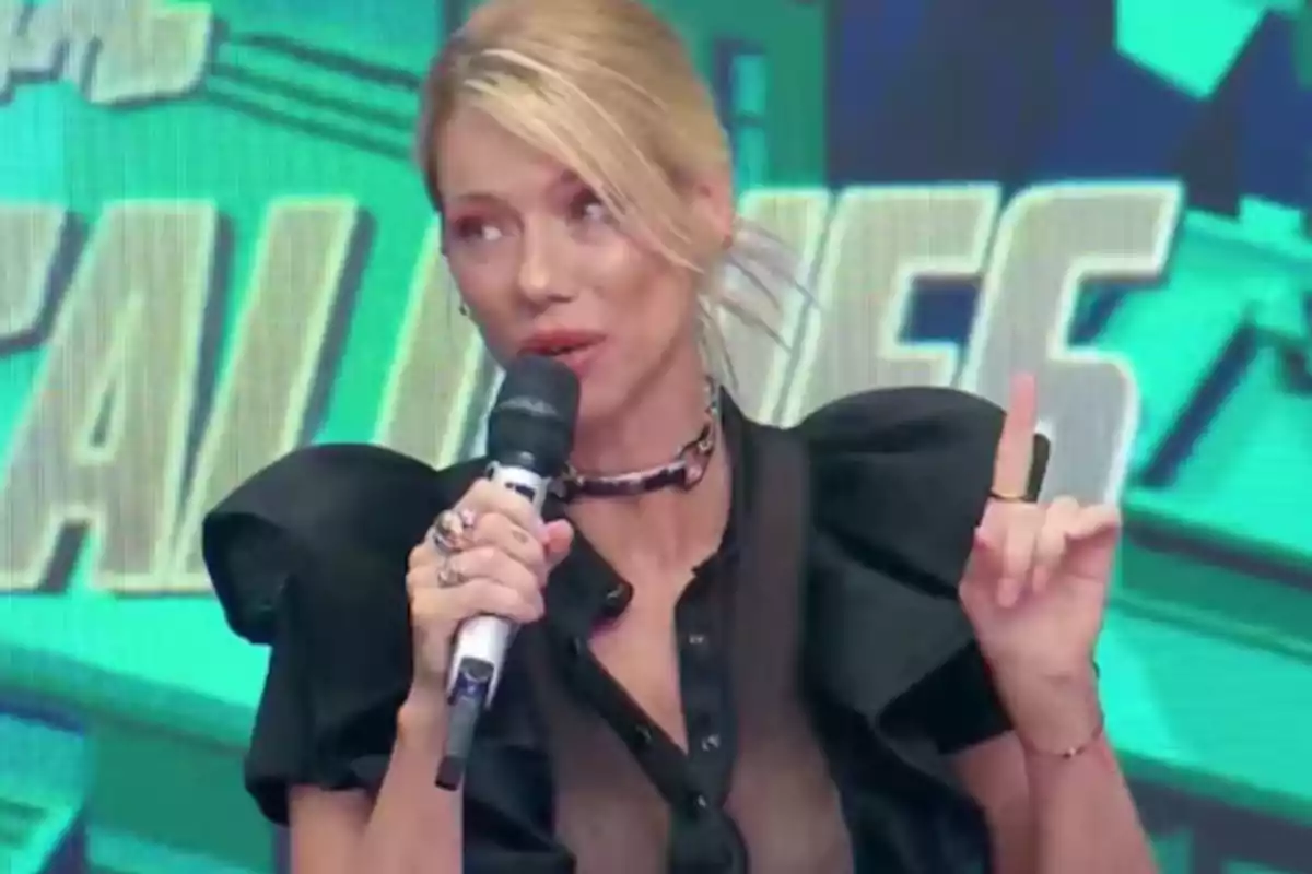 Mujer rubia con blusa negra transparente hablando por micrófono y levantando el dedo índice en un set de televisión con fondo verde. Mujer rubia con blusa negra transparente hablando por micrófono y levantando el dedo índice en un set de televisión con fondo verde.