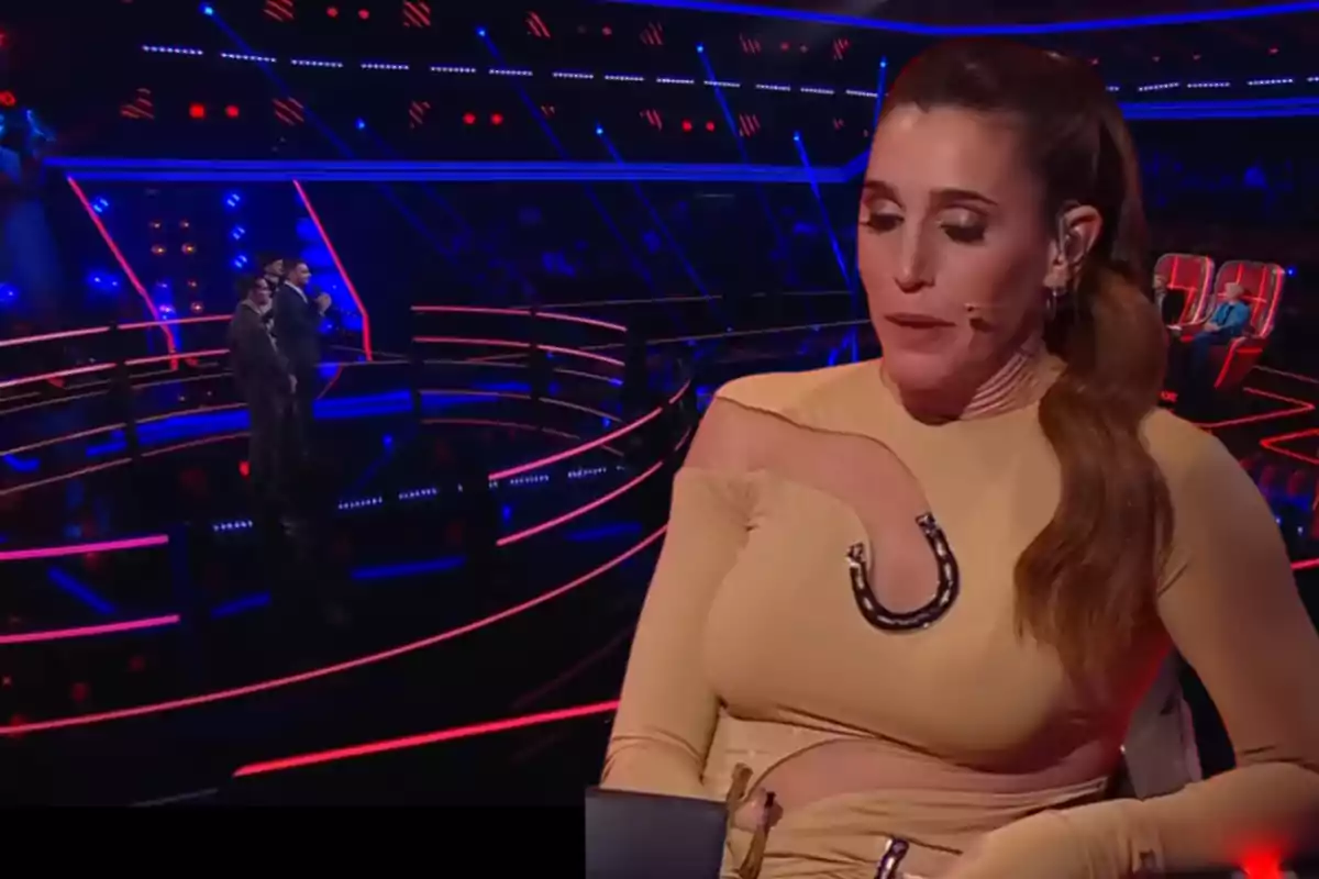 A woman in a beige dress with a headset microphone is sitting on a television set with red and blue lights while two people are standing on the stage in the background.