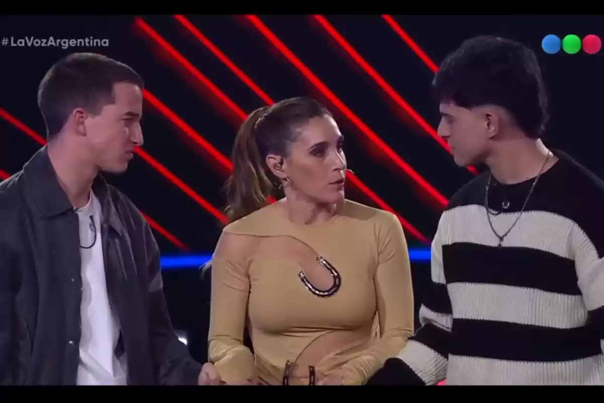 Tres personas conversando en un set de televisión con luces rojas de fondo y el logo de La Voz Argentina en la esquina superior izquierda Tres personas conversando en un set de televisión con luces rojas de fondo y el logo de La Voz Argentina en la esquina superior izquierda