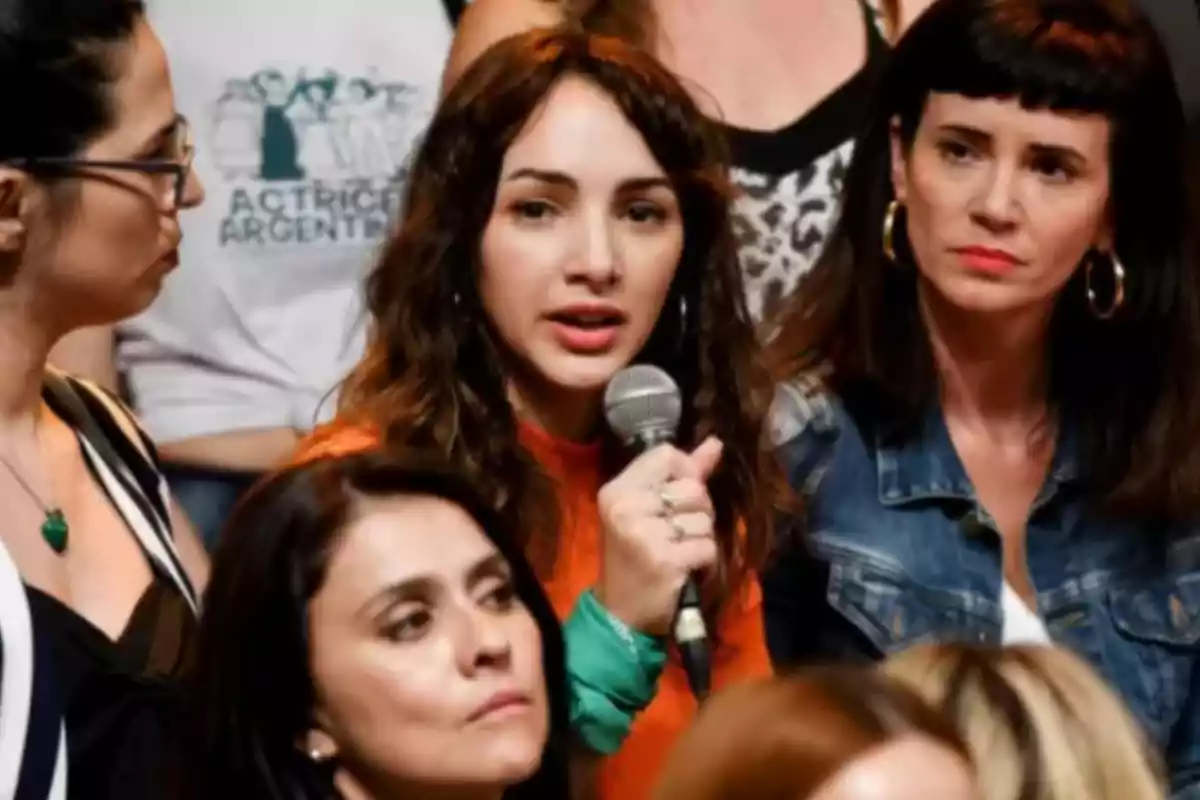 A young woman holds a microphone as she speaks among a group of women who listen alertly to her. A young woman holds a microphone as she speaks among a group of women who listen alertly to her.