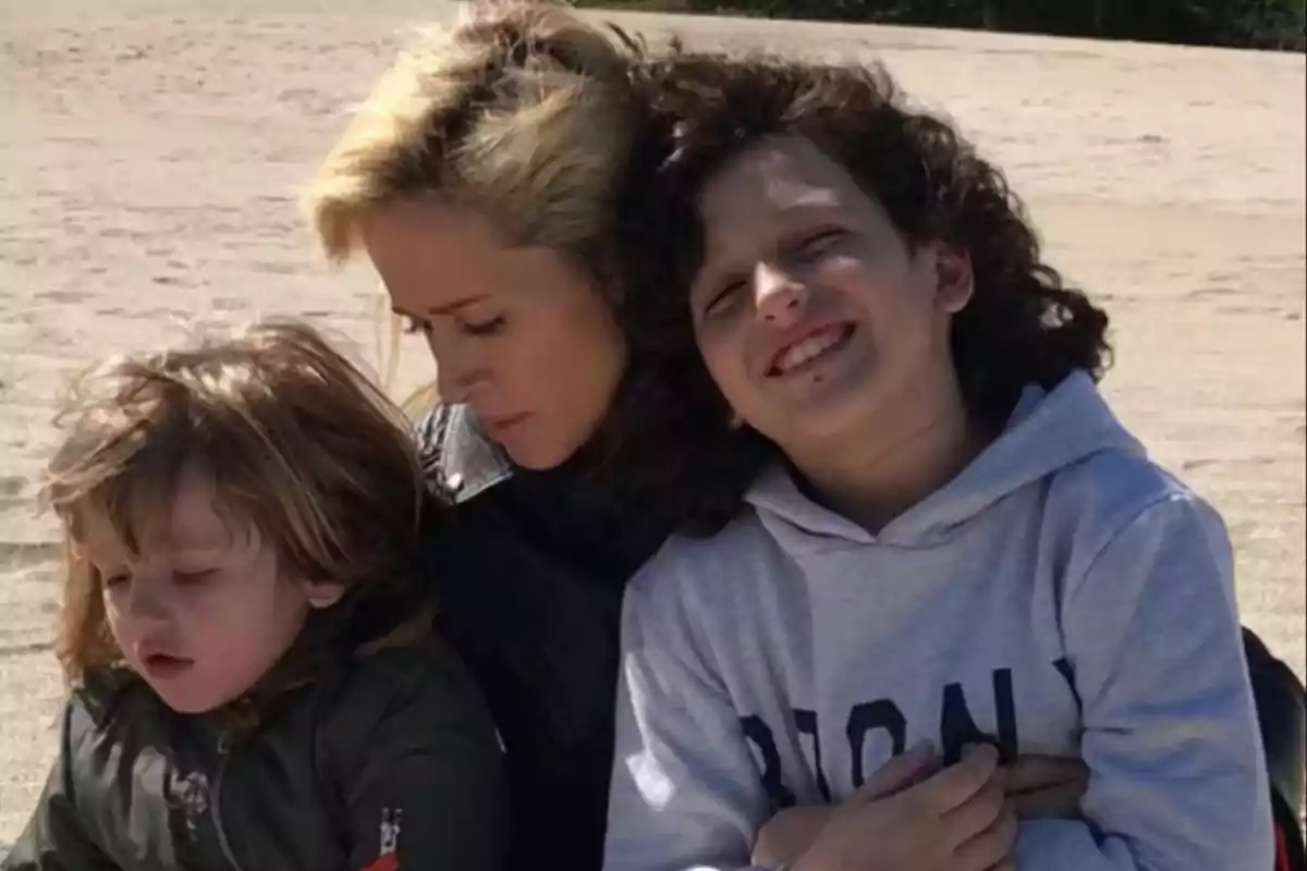 A woman hugs two small children while they sit on the sand at a beach.