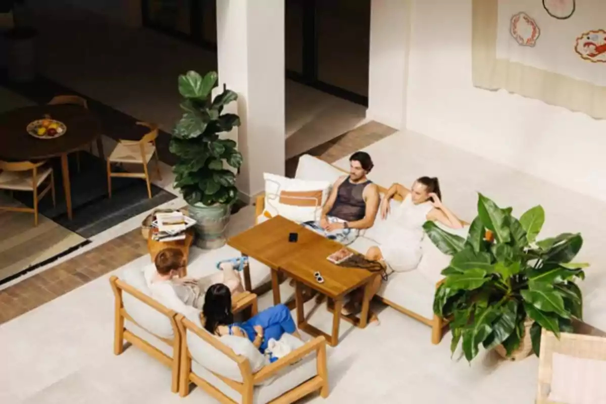 Cuatro personas conversan sentadas en una sala moderna con muebles de madera y plantas grandes Cuatro personas conversan sentadas en una sala moderna con muebles de madera y plantas grandes