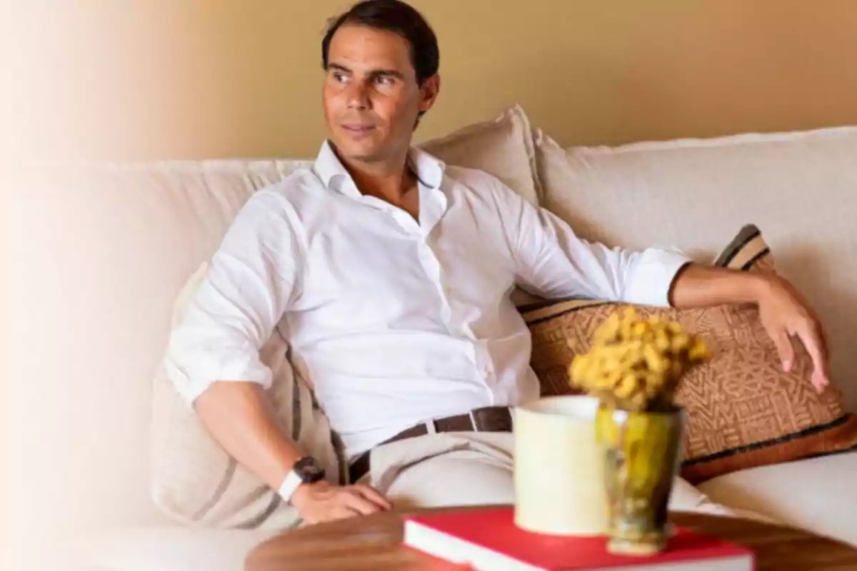 Hombre sentado en un sofá claro con camisa blanca mirando hacia un lado y una mesa con libros y un jarrón en primer plano Hombre sentado en un sofá claro con camisa blanca mirando hacia un lado y una mesa con libros y un jarrón en primer plano
