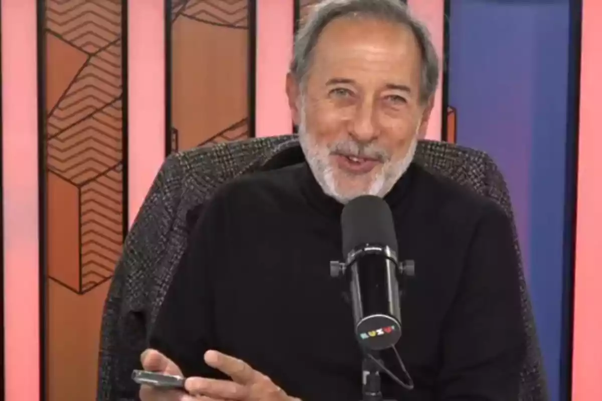 Gray-haired, bearded man sitting in front of a microphone with a colorful background, smiling while holding a mobile phone Gray-haired, bearded man sitting in front of a microphone with a colorful background, smiling while holding a mobile phone