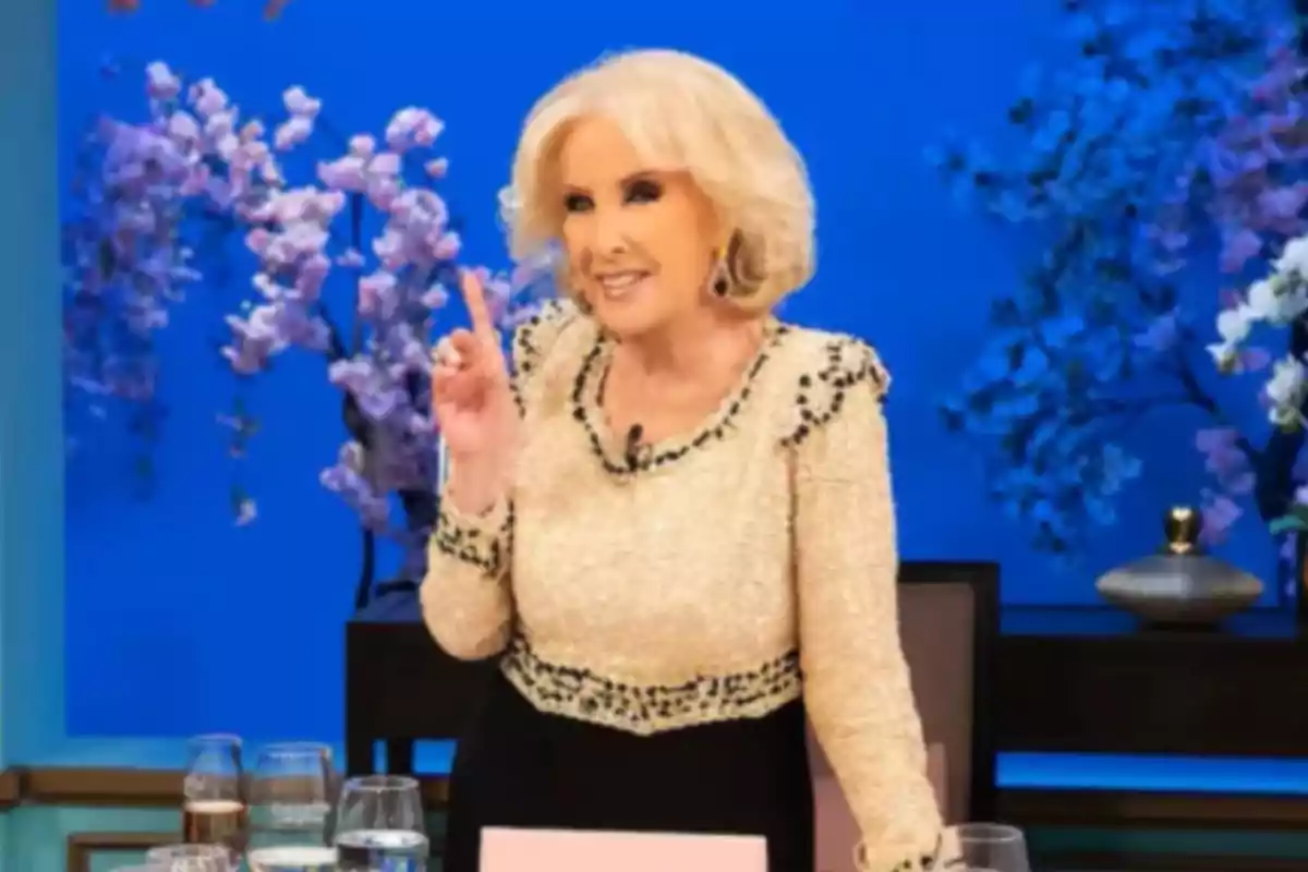 Mujer de cabello rubio y corto vestida elegantemente sonríe y levanta un dedo mientras está de pie junto a una mesa con copas y un fondo azul decorado con flores