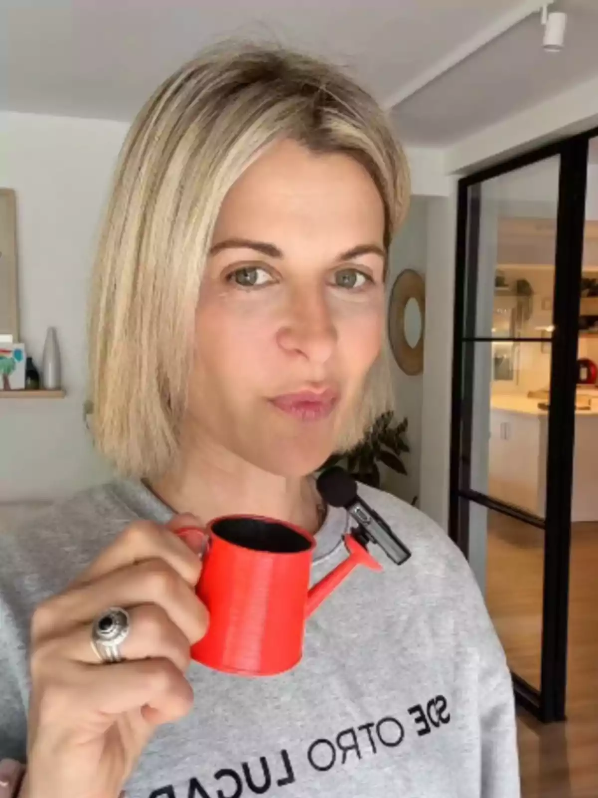 Mujer rubia con cabello corto sostiene una pequeña regadera roja mientras mira a la cámara en una habitación iluminada Mujer rubia con cabello corto sostiene una pequeña regadera roja mientras mira a la cámara en una habitación iluminada