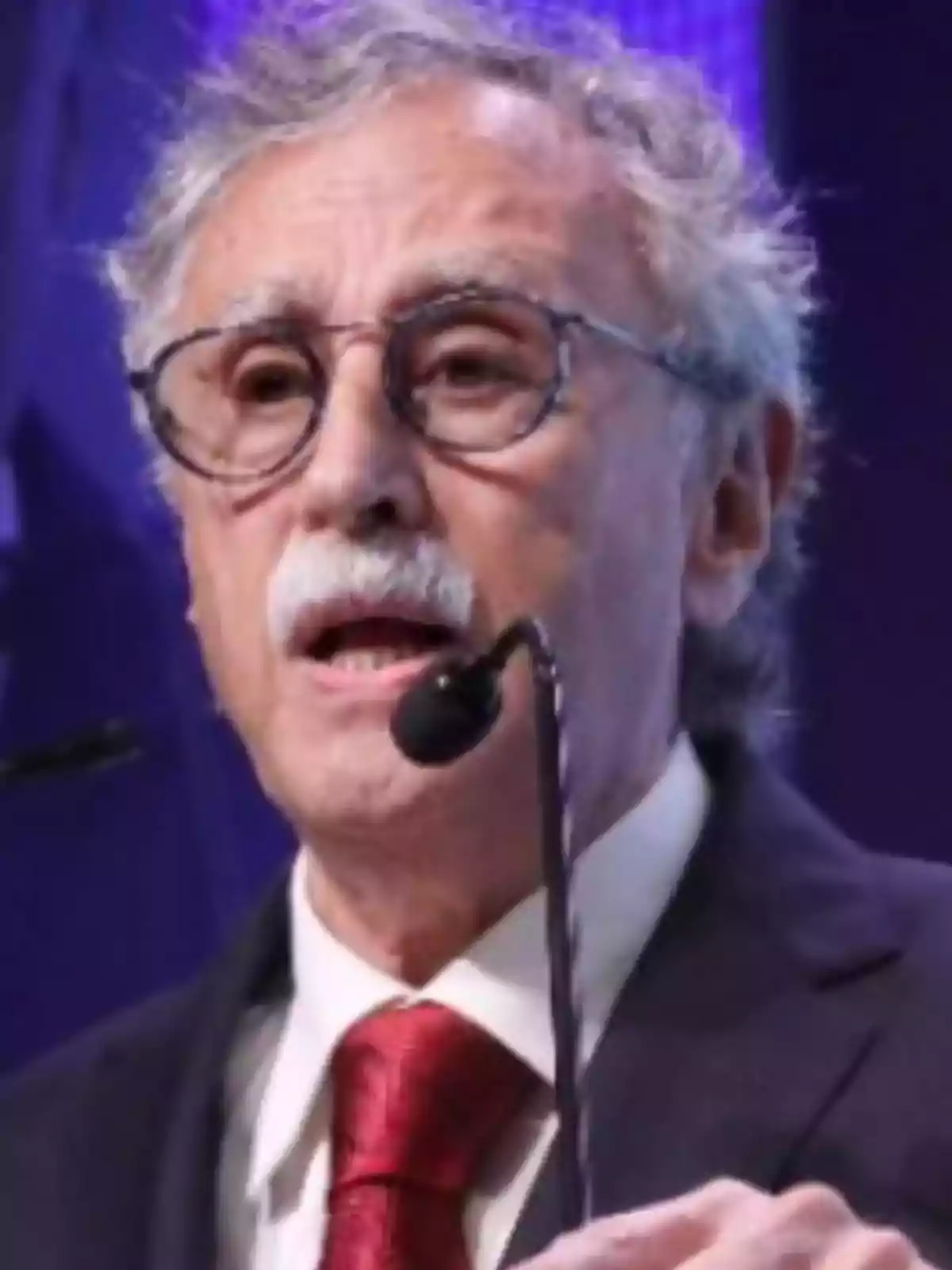Older man with glasses and a graying mustache speaking in front of a microphone while wearing a dark suit and a red tie Older man with glasses and a graying mustache speaking in front of a microphone while wearing a dark suit and a red tie