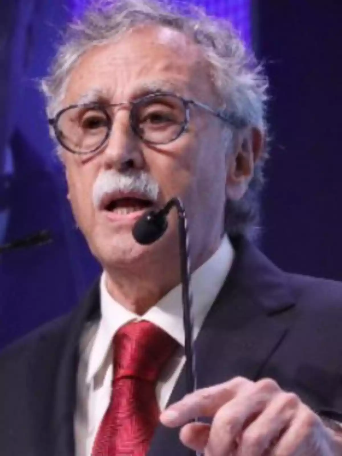 Older man with glasses and a mustache speaking into a microphone while wearing a dark suit and a red tie Older man with glasses and a mustache speaking into a microphone while wearing a dark suit and a red tie