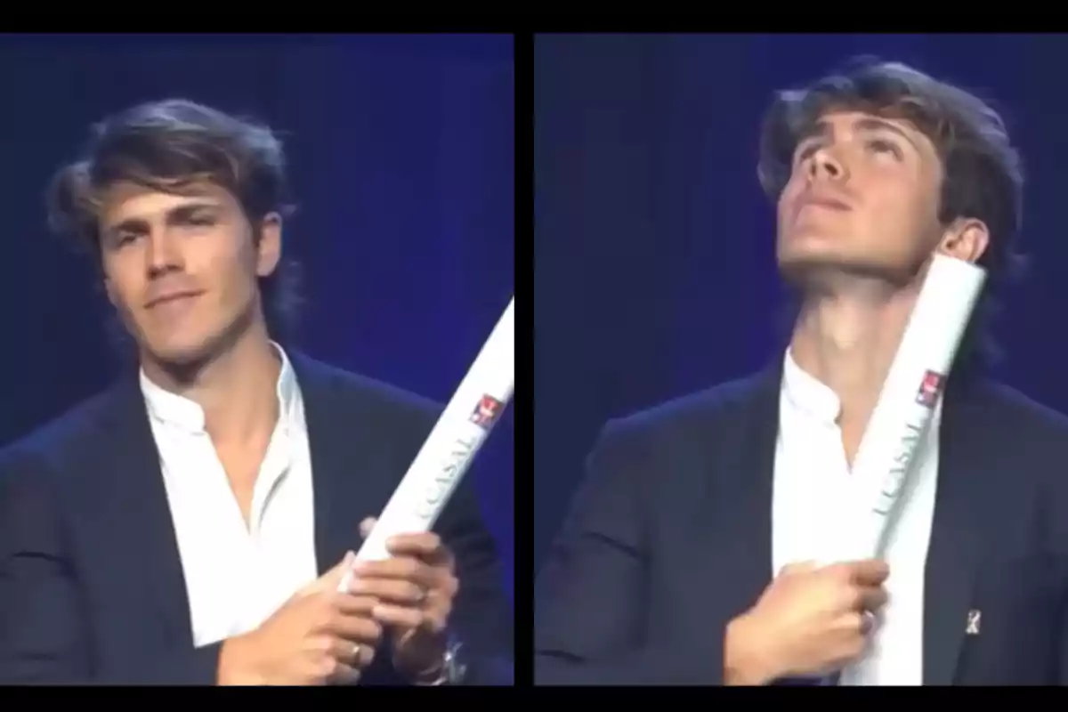 Joven con traje oscuro y camisa blanca sostiene un diploma enrollado mientras sonríe y mira hacia arriba en un escenario iluminado con fondo azul Joven con traje oscuro y camisa blanca sostiene un diploma enrollado mientras sonríe y mira hacia arriba en un escenario iluminado con fondo azul