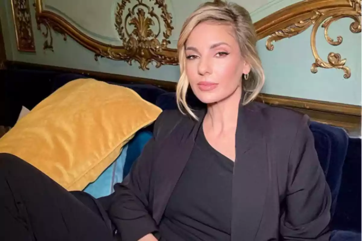 Mujer rubia con saco negro sentada en un sofá azul con cojines amarillos y fondo decorado elegantemente Mujer rubia con saco negro sentada en un sofá azul con cojines amarillos y fondo decorado elegantemente
