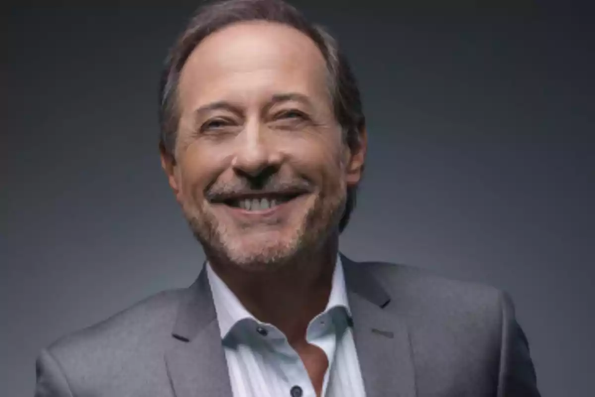 Hombre de mediana edad con barba y cabello corto sonríe mientras viste un saco gris y camisa blanca sobre un fondo oscuro. Hombre de mediana edad con barba y cabello corto sonríe mientras viste un saco gris y camisa blanca sobre un fondo oscuro.