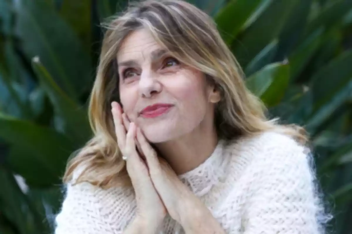 Mujer de cabello rubio con suéter blanco apoyando el rostro en sus manos y mirando hacia arriba con expresión pensativa, fondo de plantas verdes desenfocado