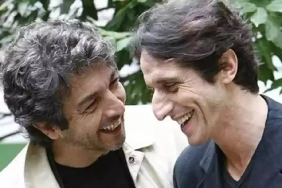Dos hombres sonríen y se miran con complicidad en un entorno al aire libre con plantas de fondo