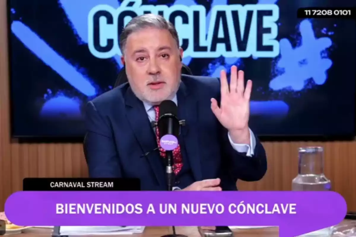 Un hombre de traje habla frente a un micrófono en un estudio con un letrero que dice bienvenidos a un nuevo cónclave