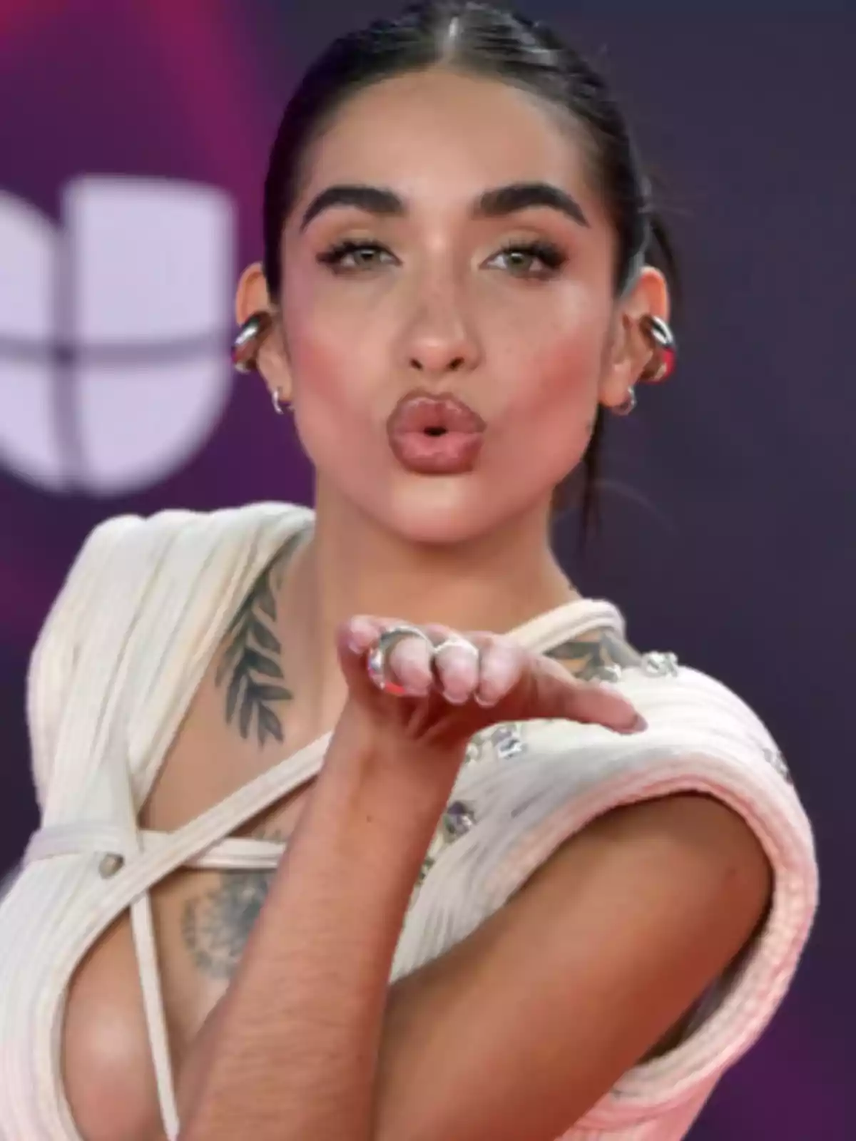 Mujer joven con cabello oscuro recogido, vestida de blanco, lanzando un beso con la mano frente a un fondo desenfocado