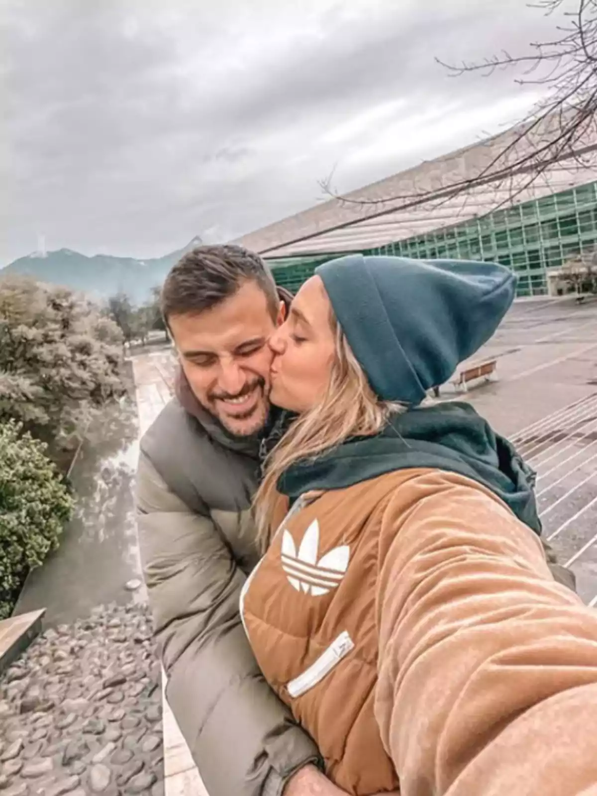 Pareja abrazada tomándose una selfie mientras la mujer besa al hombre en la mejilla en un entorno al aire libre con un edificio moderno y montañas al fondo Pareja abrazada tomándose una selfie mientras la mujer besa al hombre en la mejilla en un entorno al aire libre con un edificio moderno y montañas al fondo