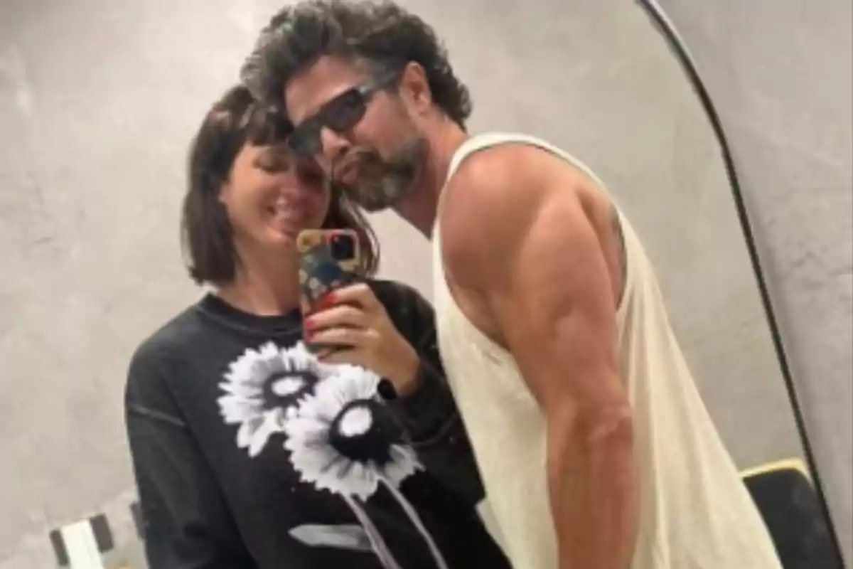 Pareja tomándose una selfie frente a un espejo en un baño, ambos sonríen y el hombre lleva gafas oscuras y camiseta sin mangas Pareja tomándose una selfie frente a un espejo en un baño, ambos sonríen y el hombre lleva gafas oscuras y camiseta sin mangas