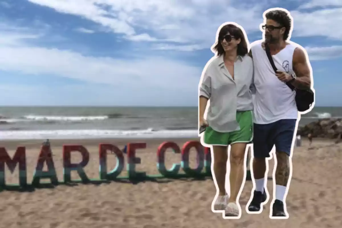 Couple walking on the beach in front of a colorful sign that says "Mar de" in the sand with the sea and sky in the background