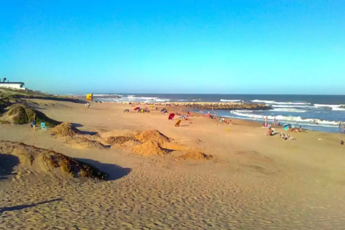 Playa amplia con arena dorada, varias personas disfrutando del sol y el mar, algunas sombrillas de colores y olas rompiendo cerca de la orilla Playa amplia con arena dorada, varias personas disfrutando del sol y el mar, algunas sombrillas de colores y olas rompiendo cerca de la orilla