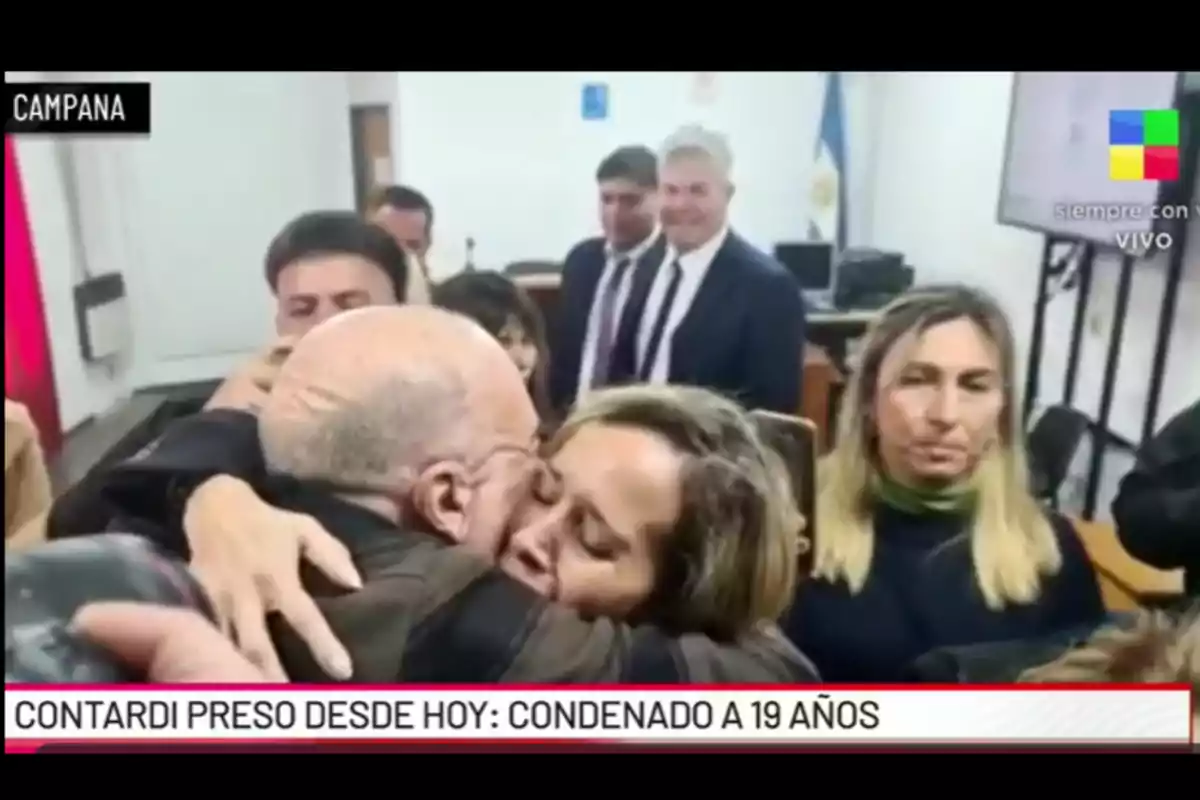 Un grupo de personas en una sala de tribunal, dos de ellas se abrazan emocionadas mientras otras observan, al fondo se ven hombres de traje y una bandera argentina. Un grupo de personas en una sala de tribunal, dos de ellas se abrazan emocionadas mientras otras observan, al fondo se ven hombres de traje y una bandera argentina.