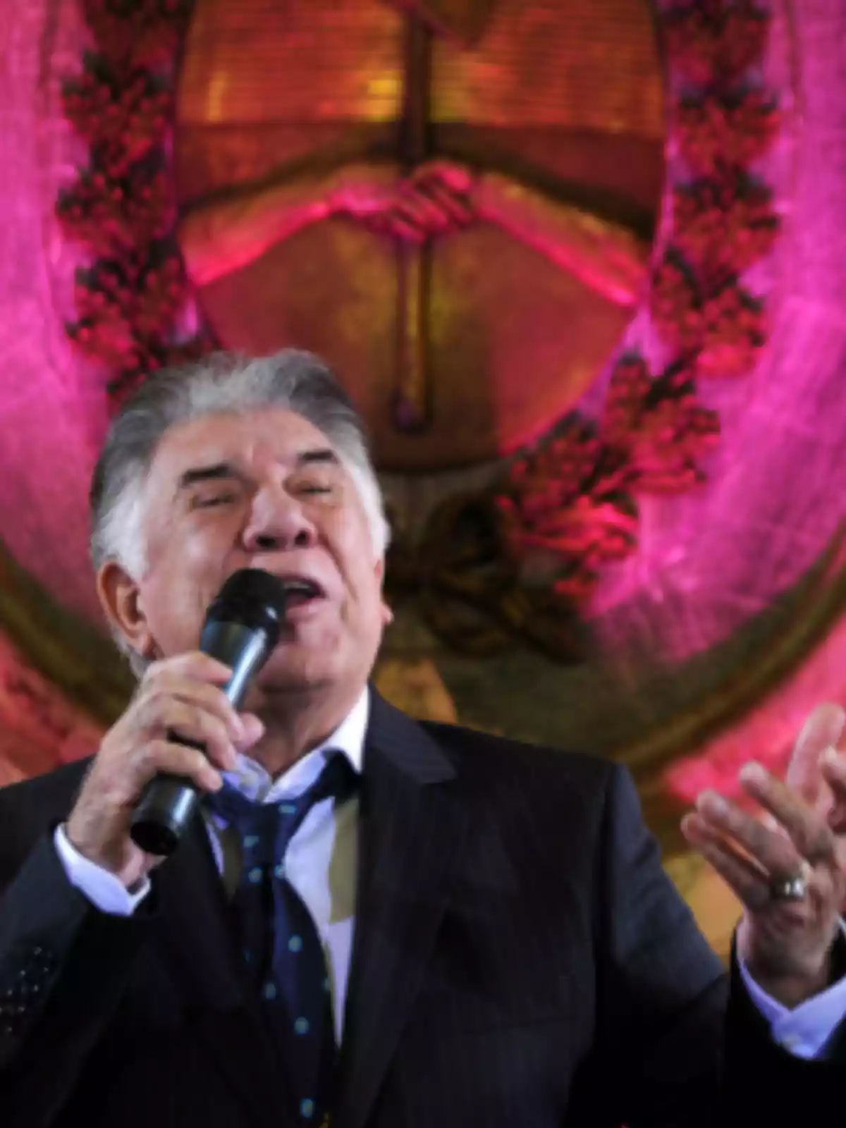 Older man with gray hair singing with a microphone in his hand, wearing a dark suit and tie, with a decorative background lit in pink and gold tones Older man with gray hair singing with a microphone in his hand, wearing a dark suit and tie, with a decorative background lit in pink and gold tones
