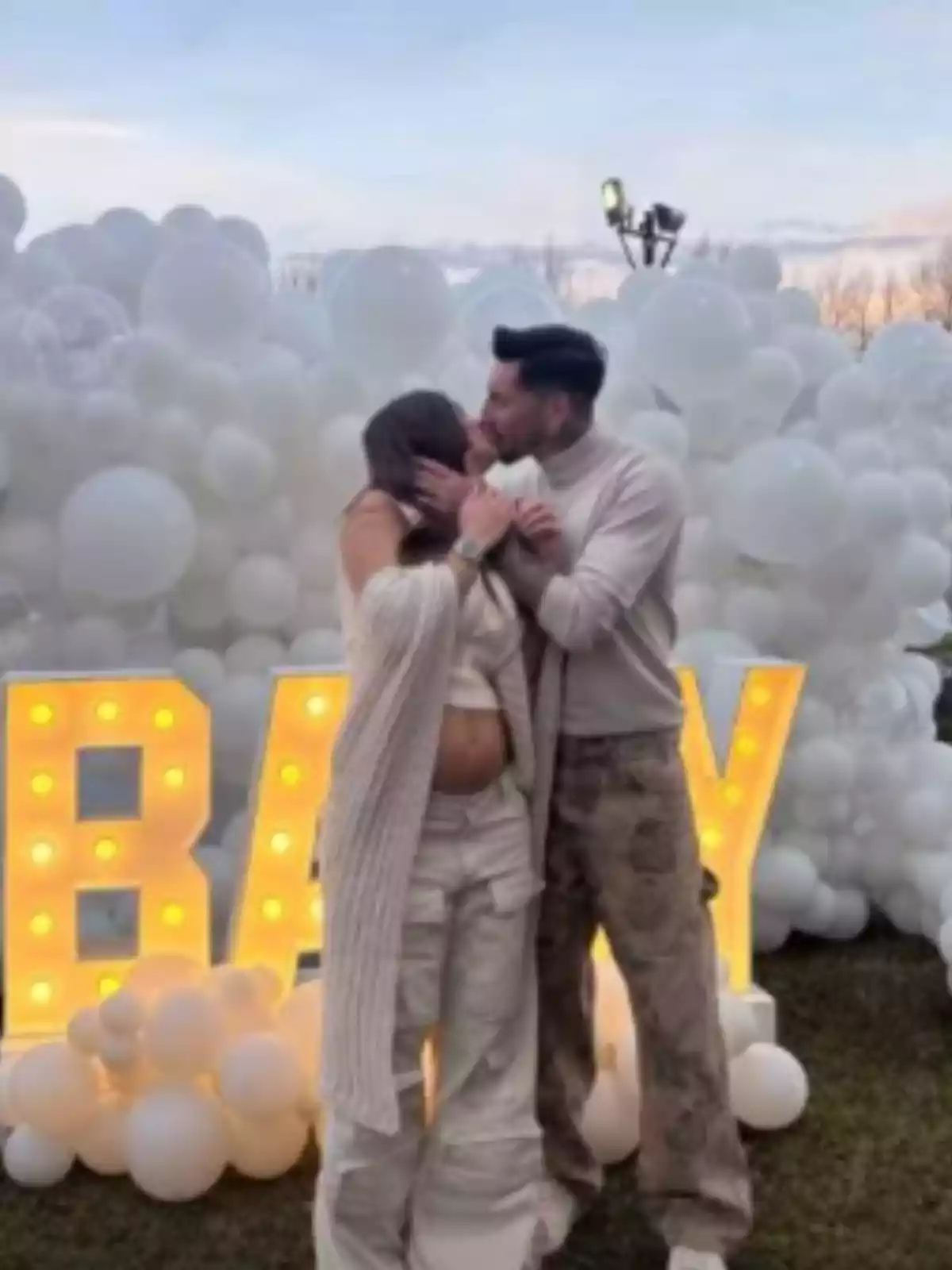 Pareja abrazándose y besándose frente a un fondo de globos blancos y letras iluminadas que forman la palabra BABY en un ambiente al aire libre Pareja abrazándose y besándose frente a un fondo de globos blancos y letras iluminadas que forman la palabra BABY en un ambiente al aire libre