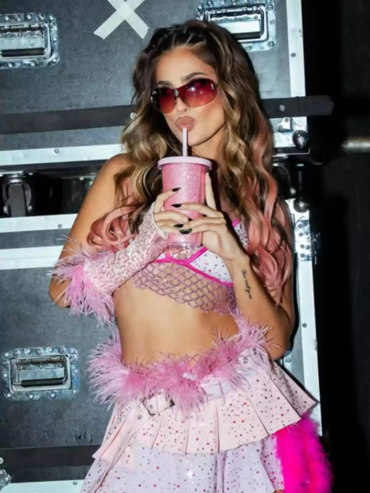 Woman with sunglasses and long wavy hair, dressed in a shiny pink outfit with feather details, holding a pink cup with a straw in front of equipment boxes
