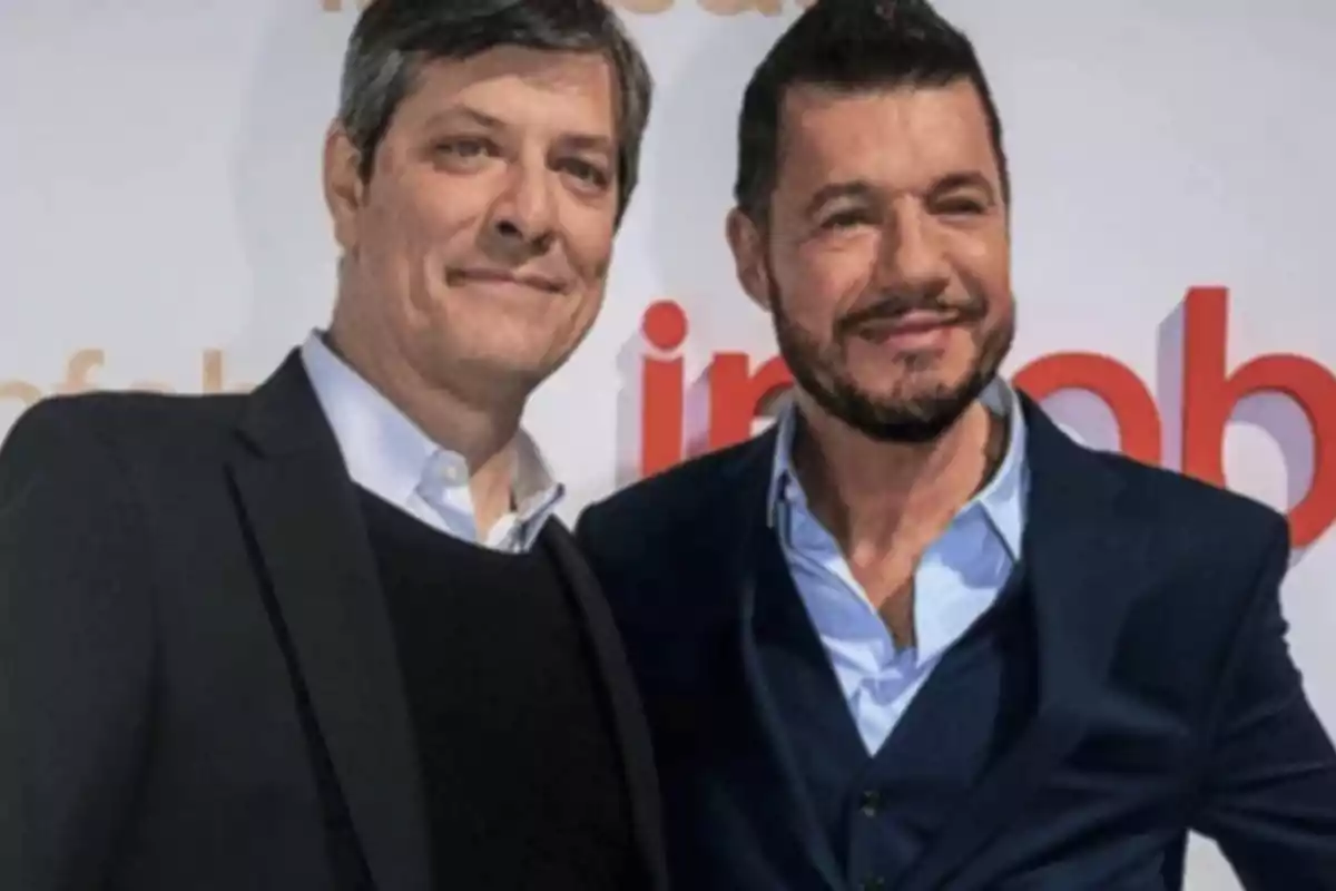 Two men dressed in suits pose together and smile in front of a background with blurred red and orange letters. Two men dressed in suits pose together and smile in front of a background with blurred red and orange letters.