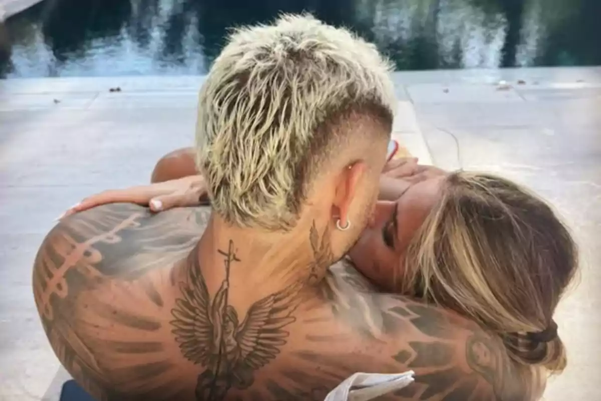 Pareja abrazada junto a una piscina, el hombre tiene el cabello rubio corto y tatuajes en la espalda, mientras la mujer apoya su cabeza en su hombro Pareja abrazada junto a una piscina, el hombre tiene el cabello rubio corto y tatuajes en la espalda, mientras la mujer apoya su cabeza en su hombro